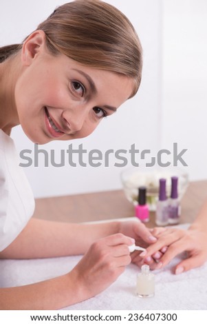 manicurist doing manicure to  client  looking at camera painting nails with transparent nail polish  in salon  on white towel