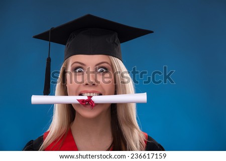 Portrait of beautiful happy blond girl in black cloak and master hat holding in mouth bundle of sheet of paper on blue background smiling looking at camera