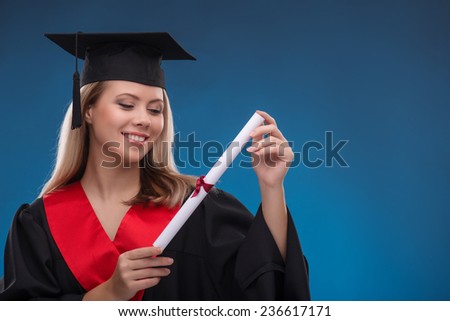 Portrait of beautiful happy blond girl in black cloak and master hat looking at bundle of sheet of paper on blue background smiling with copy place