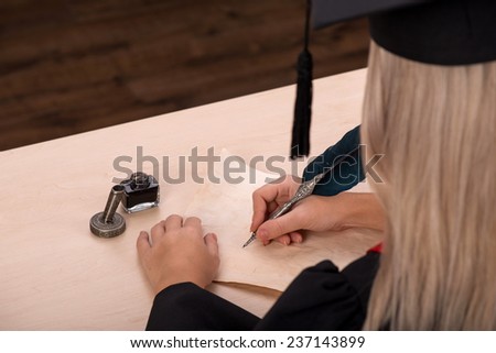 Portrait of beautiful happy blond girl in black cloak and master hat writing with ink and feather