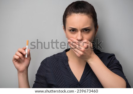 Closeup of young attractive Caucasian woman determined girl holding cigarette and shutting mouth with hand. Quit smoking. Studio shot isolated on grey. Addiction concept