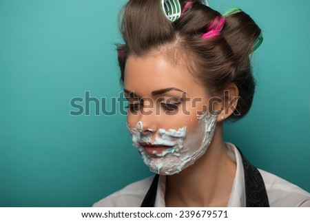 Cute brunette woman in hair curlers playing fool with foam on face looking down, isolated on blue background, role gender reversal concept