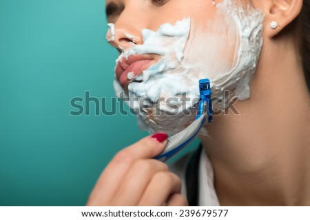 Closeup portrait of brunette shaving her face with foam and razor isolated on blue background. Role gender reversal concept