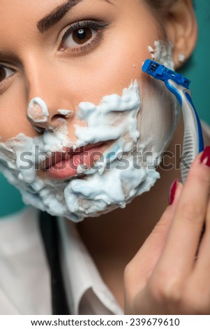 Closeup portrait of brunette shaving her face with foam and razor isolated on blue background. Role gender reversal concept