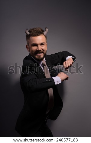 Business games.  Devilish cunning businessman with horns on head in full suit dancing and smiling happily against grey background