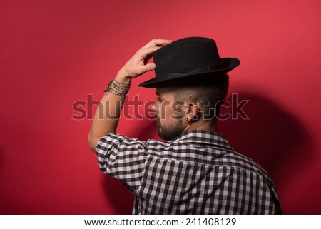Handsome in hat. Young stylish man holding hands on hat and looking from behind while standing against red background