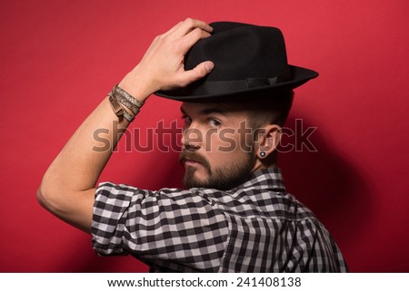 Handsome in hat. Young stylish man holding hands on hat and looking from behind while standing against red background