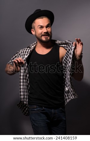 Having fun. Cheerful handsome young man in hat posing pulling his shirt in a funny manner while standing against grey background