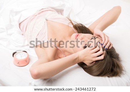 Too much noise. Young beauty lying in bed holding hands on head with a clock on blanket against white background