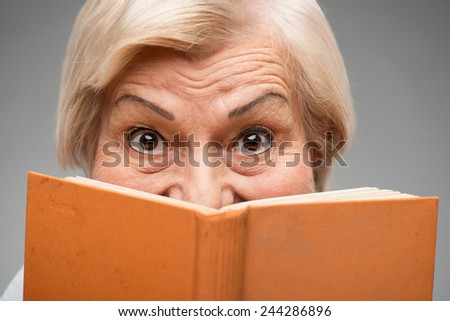 I love reading. Closeup of smiling senior woman holding orange book hiding her face behind it while standing against grey background
