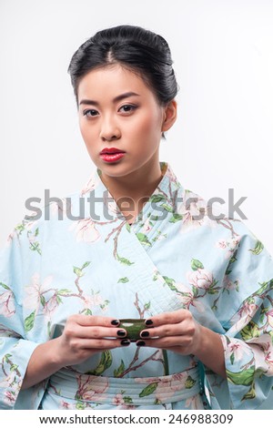Asian beauty enjoying tea ceremony. Young beautiful Asian woman in traditional kimono holding cup of tea and looking at camera while standing against white background