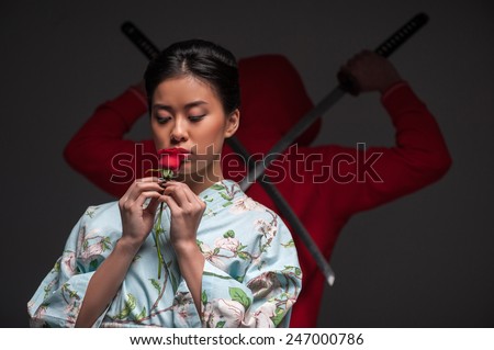 Oriental culture and traditions. Young beautiful Japanese woman in kimono holding a rose while standing against grey background with traditional ninja warrior with two swords
