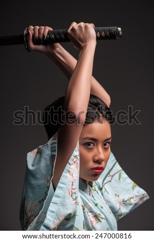 Aggressive move. Side view portrait of young beautiful Japanese woman in kimono attacking with katana sword while standing against grey background