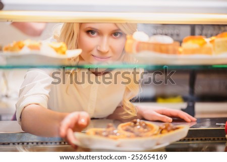 Please enjoy. Closeup image of beautiful young female baker stretching out two paper cups while standing against cafe with selective focus