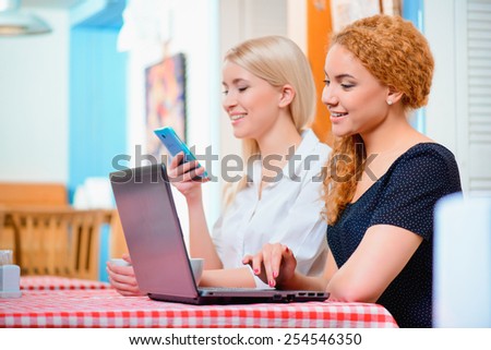No minute without laptop. Side view of two young women working on laptop and looking at mobile phone while sitting on the couch of the cafe