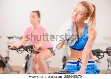 Cycling on exercise bikes. Two attractive young women in sports clothing exercising on gym bicycles while blond girl is wiping her neck with a towel