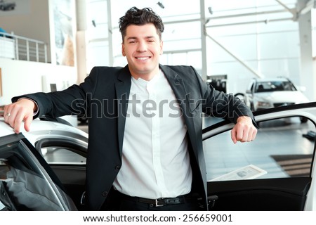 Confident in his choice. Smiling handsome man in suit standing by a new car and smiling at camera in car dealership