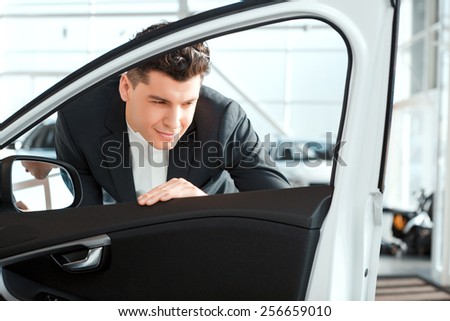 Love luxury cars. Smiling handsome man in formalwear looking inside the new car in car dealership
