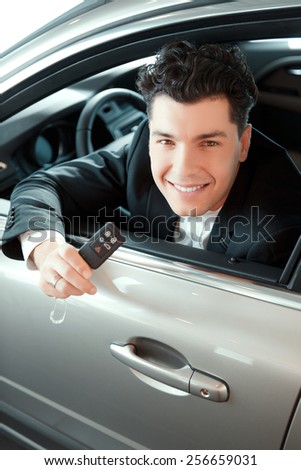 Happy car owner. Portrait of a smiling handsome man in suit sitting on drivers place in car and looking through the car window stretching out the key in car dealership
