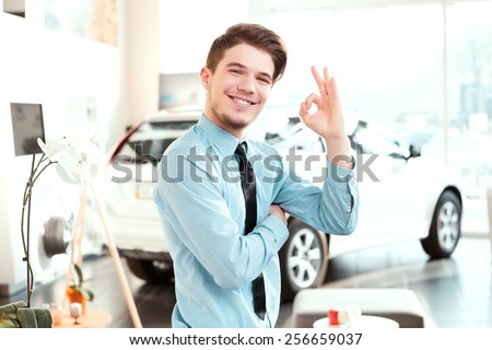It is perfect choice. Portrait of a handsome young car sales man in formalwear showing ok sign and smiling at camera in a car dealership