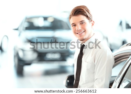 I love my job. Portrait of a handsome young car sales man in formalwear leaning at car and looking at camera in a car dealership