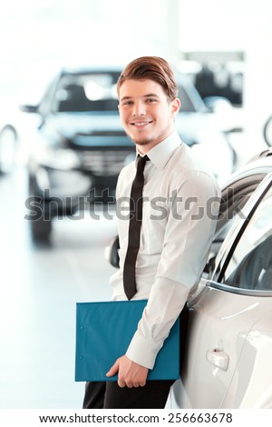 Assistant in vehicle search. Portrait of a handsome young car sales man in formalwear holding a clipboard and looking at camera in a car dealership