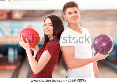 We are a team. Cheerful young man and women posing back to back while standing against bowling alleys and holding balls