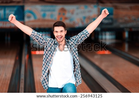 Bowling master. Cheerful young man raising his hands high above the head and smiling at camera while standing against bowling alleys after the strike