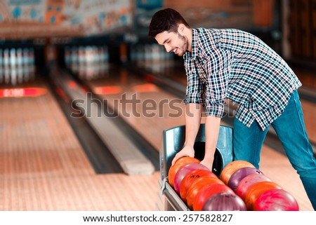 Choosing the lucky ball. Cheerful young man choosing bowling ball and smiling while standing against bowling alleys