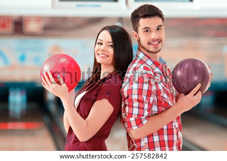 We are a team. Cheerful young man and women posing back to back while standing against bowling alleys and holding balls