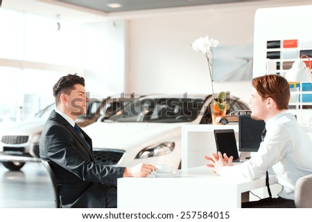 Discussing the deal. Side view of a young handsome salesman speaking with his satisfied client in car dealership while sitting at the table and drinking coffee