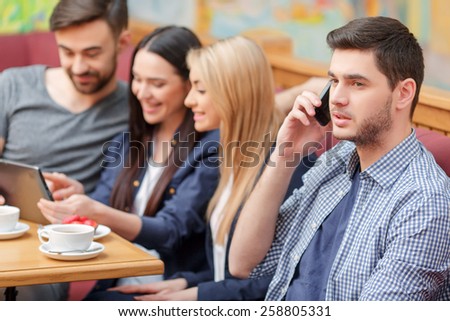 Staying always connected. Handsome young man in casual clothing talking on his mobile phone while witting in the cafe with his friends on the background surfing in the web with digital tablet