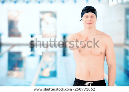Born to swim.  Young handsome man with naked torso wearing a swimming cap and goggles while standing in the swimming pool and showing this thumb up