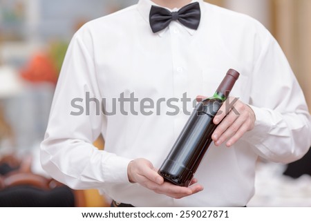 Confident waiter. Cropped image of handsome young waiter in shirt and bow tie holding a wine bottle in the luxury restaurant