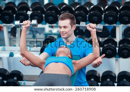 Attentive sports instructor. Beautiful young woman in sports clothing exercising with dumbbells while lying on the bench of gym under control of her sports instructor