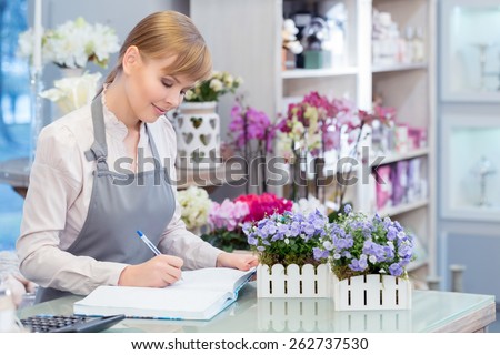 Growing her flower business. Young attractive florist making notes at her note pad sitting by the counter of the flower shop
