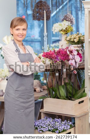 Proud of her job. Female florist in grey apron standing by the stand with floral wooden boxes