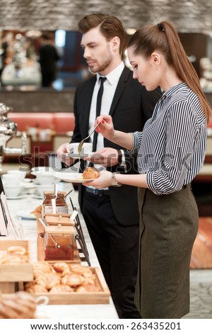 Something sweet for the lunch. Attractive young businesswoman taking jam to add to her croissant on the plate with her male colleague on the background