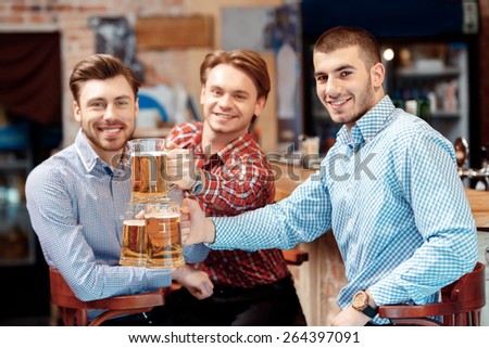 Sharing beer with good friend. Three cheerful young stretching out glasses with beer looking at the camera