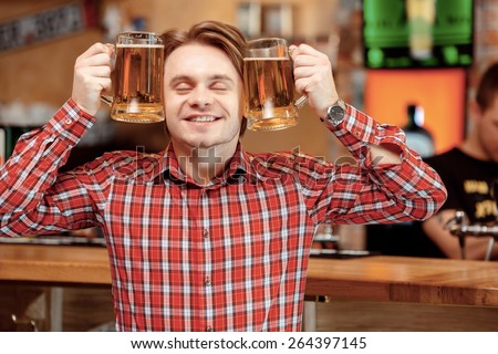 Happy together with friends. Young cheerful man holding two mugs with beer by his face and smiling with closed eyes