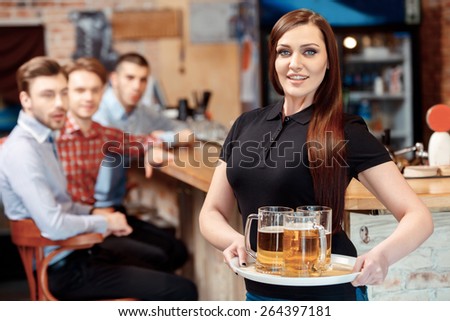What a beautiful girl. Selective focus on beautiful waitress holding a tray with beer glasses while young men are looking at her with amazement