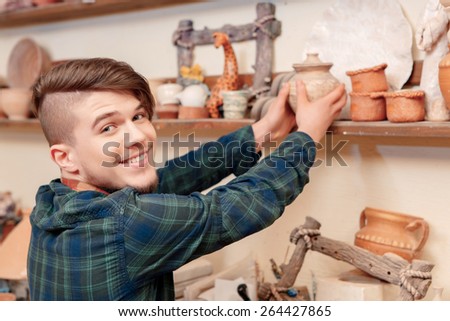 Happy in art. Young handsome guy putting a clay jar on the shelf smiling to the camera