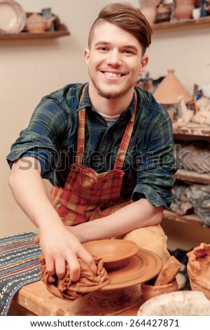 Joyful job. Young male potter shaping clay pot in pottery wheel and smiling to the camera
