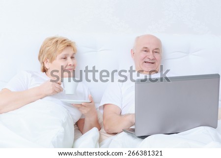 Socializing in bed. Elderly female drinking tea with her husband using laptop lying in bed