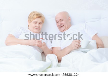 Message from the family. Elderly couple smiling and excited reads text message on the phone lying in bed