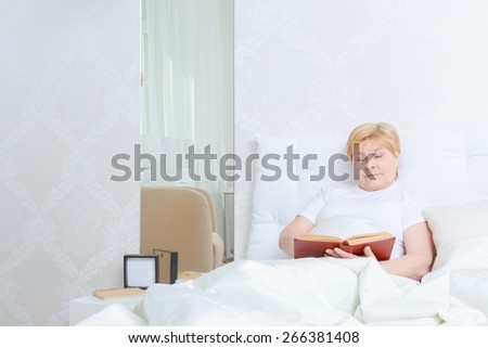 Thoughtful reading. Elderly woman in white t-shirt reading a book in bed before going to sleep
