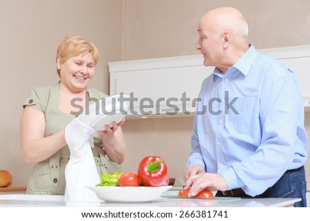 Family time. Cute elderly couple at the kitchen where husband is cutting vegetables and talking to his wife drying dishes and smiling