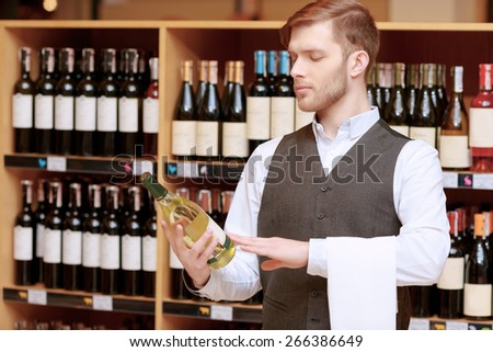 Expert in wine. Young sommelier looking seriously at the label of a wine bottle and holding a towel on his hand
