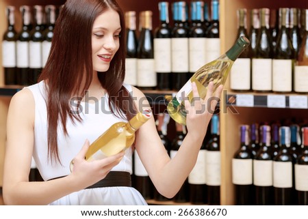 Making choice. Young smiling woman with expression of hesitation comparing two bottles of wine in a liquor store