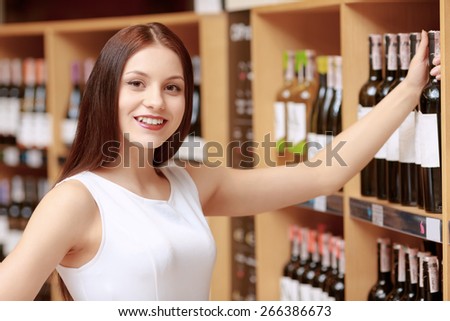 Light citrus wine. Close-up of a young smiling woman taking a wine bottle from the store shelf and smiling to the camera satisfied with her choice